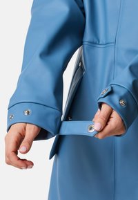 Niebieski wodoodporny płaszcz o gładkiej teksturze. Posiada mankiety zapinane na zatrzask oraz boczną kieszeń. Tkanina ma matowe wykończenie.