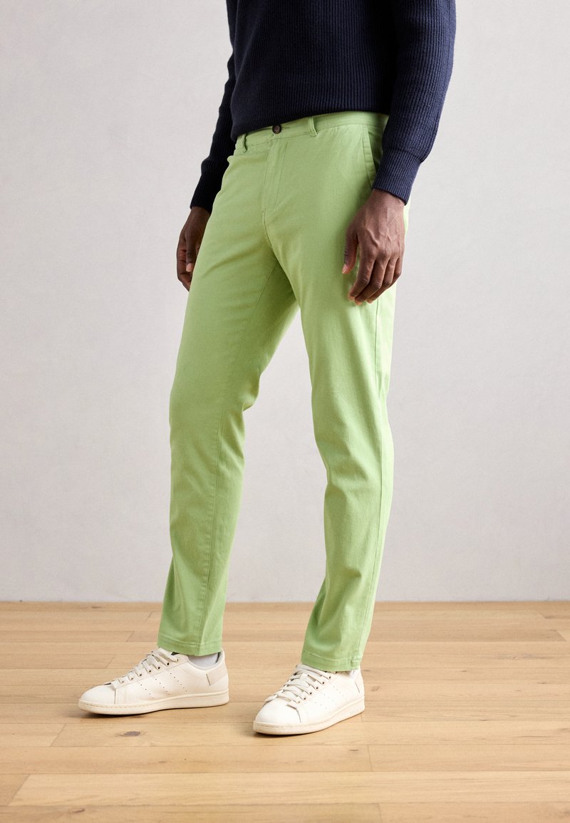 Chino in cotone verde chiaro con vestibilità slim, tasche frontali e chiusura a bottone, abbinati a sneakers bianche, su un pavimento in legno.