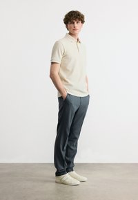 Νεαρός άνδρας όρθιος με τα χέρια στις τσέπες, φοράει κρεμ υφασμάτινη πόλο μπλούζα, σκούρο γκρι παντελόνι και λευκά αθλητικά παπούτσια, σε απλό φόντο.