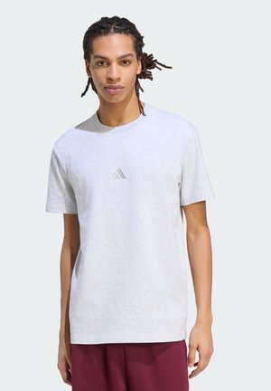 Giovane uomo in piedi, indossa una maglietta Adidas grigio chiaro e pantaloni bordeaux, con i capelli intrecciati legati dietro e un'espressione neutra.