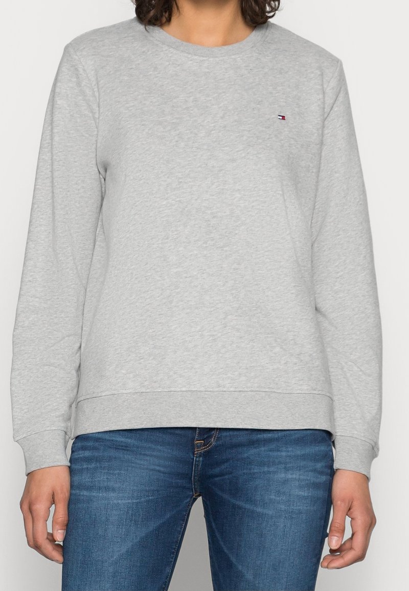 Personne portant un sweat-shirt gris clair uni à manches longues avec un petit logo sur la poitrine et un jean bleu en denim.