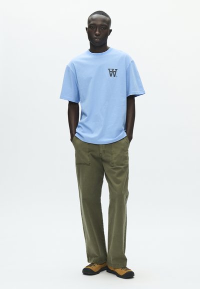 T-shirt en coton bleu clair avec un logo noir sur la poitrine, associé à un pantalon vert olive. Tenue complétée par des chaussures marron et noires.