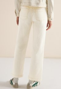 Personne portant un jean large couleur écru, une veste assortie, des chaussettes grises et des baskets blanches à rayures vertes, debout devant un fond beige.