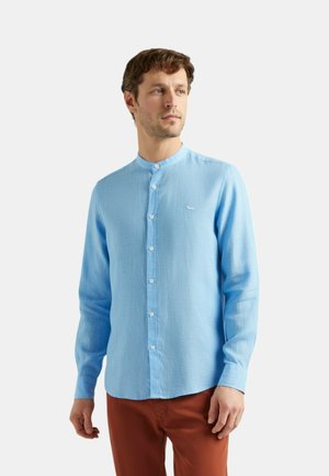 Uomo che indossa una camicia celeste con colletto alla coreana e pantaloni color ruggine, in piedi su sfondo bianco, guarda verso destra.