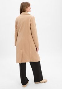Beige Wollmischungsjacke mit einem maßgeschneiderten Kragen, Rückenschlitz und langen Ärmeln, kombiniert mit schwarzen weit geschnittenen Hosen und beigen Plateauschuhen.
