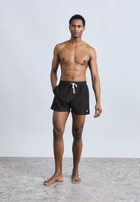 Μαύρα μαγιό σορτς με ελαστική μέση και λευκό κορδόνι, με πλαϊνές τσέπες και μικρό λογότυπο στο τελείωμα.