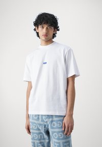 White cotton t-shirt with a crew neck and relaxed fit, featuring a small blue label on the chest. Worn with patterned light blue jeans.