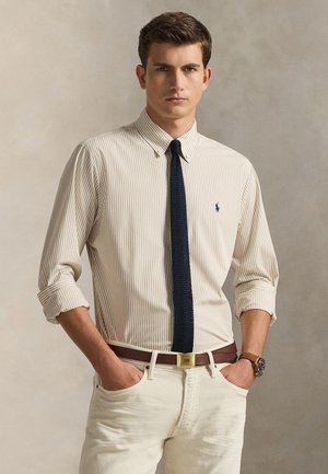 Camicia a righe beige e crema con bottoni, abbinata a una cravatta blu navy, cintura marrone e pantaloni chiari. Maniche arrotolate.