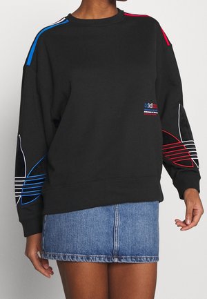 Czarna oversize bluza z niebieskimi i czerwonymi paskami na ramionach i rękawach, małe wielokolorowe logo na klatce piersiowej, noszona z dżinsową spódnicą.