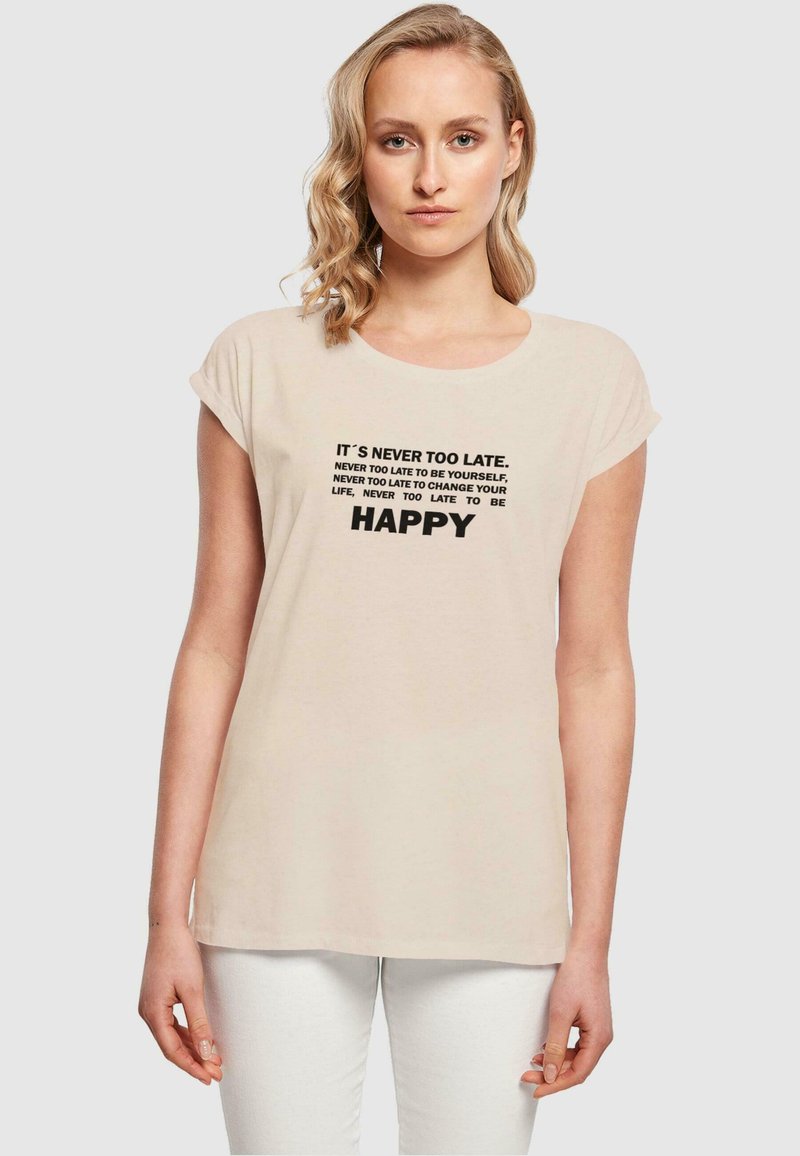 Helle beige T-Shirt mit kurzen Ärmeln und rundem Halsausschnitt. Aufgedruckter Text in Schwarz: motivierende Zitate, darunter "ES IST NIE ZU SPÄT" und "GLÜCKLICH."