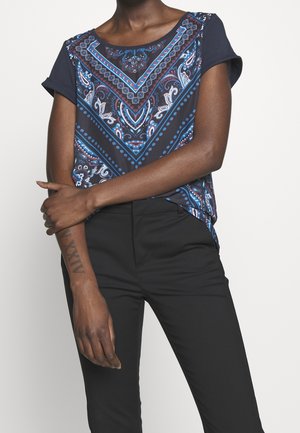 Personne portant un haut à manches courtes bleu marine à motifs avec des accents rouges et blancs et un pantalon noir, le bras droit croisé sur le corps.