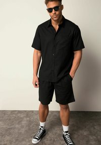 Camicia nera a maniche corte con bottoni e tasca, abbinata a pantaloni corti neri coordinati. Il modello indossa occhiali da sole e sneaker nere alte.