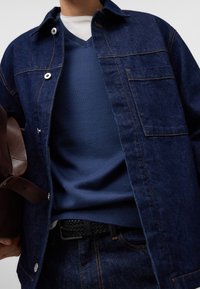 Dunkelblaue Jeansjacke mit silbernen Knöpfen, über einem marineblauen V-Ausschnitt-Pullover getragen. Abgerundet mit einem geflochtenen Leder-Gürtel und einer braunen Tasche.