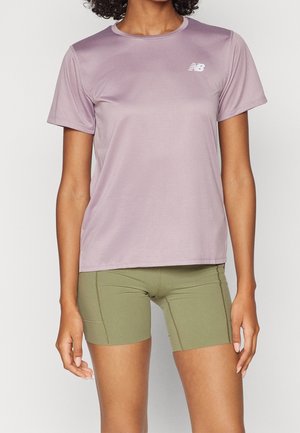 Mujer que lleva una camiseta deportiva de manga corta de color lila claro y pantalones cortos ajustados de color verde oliva, de pie frente a un fondo liso.