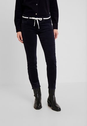 Mujer de pie con pantalones negros ajustados con cinturón blanco de cordón y botines negros con tacón, sobre un fondo claro y liso.