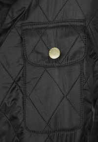 Tecidos acolchoados pretos com um bolso de remendo quadrado e fechamento com botão de pressão dourado, detalhado com costura em diamante e textura suave.