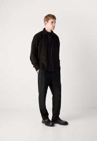 Black corduroy shirt over a dark top, paired with black trousers and glossy black shoes. Model stands against a light gray background.