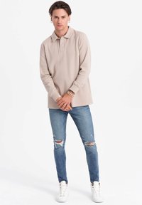 Beige långärmad polo-tröja med krage, kombinerad med blå slitna skinny jeans och vita hög-top sneakers. Avslappnad och figursydd design.