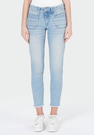 Jean slim en denim bleu clair avec poches avant et ourlets effilochés, porté avec des baskets blanches et un haut blanc.