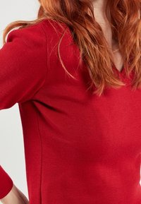 Rotes gestricktes Oberteil mit V-Ausschnitt und taillierten Ärmeln, mit einer glatten Textur und gerippten Details entlang der Nähte.