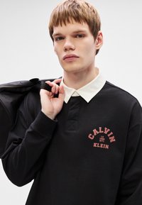 Μαύρο πουκάμισο πόλο με μακριά μανίκια και λευκό γιακά, που διαθέτει μια κόκκινη γραφική λογότυπο που γράφει "CALVIN KLEIN" στο στήθος. Η υφή είναι λεία.