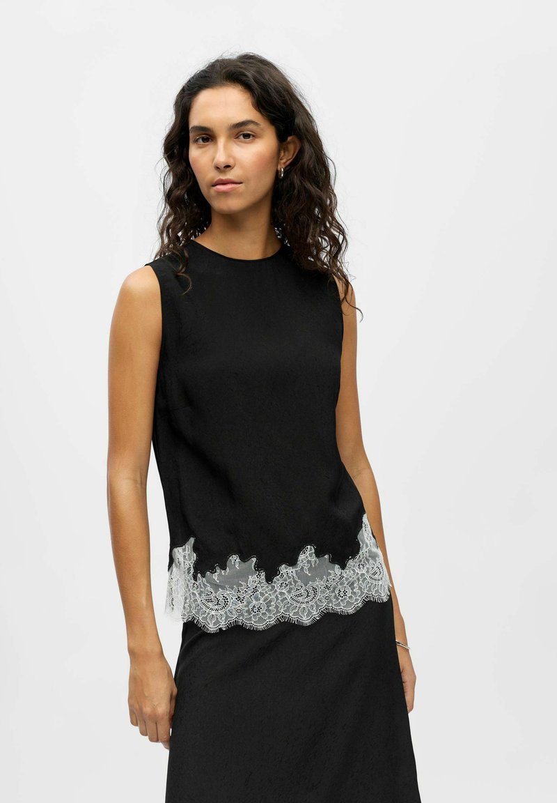 Haut noir sans manches en tissu texturé avec une bordure en dentelle à l'ourlet ornée de motifs floraux. Aucun fermoir ni accent visible.