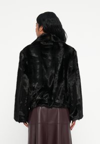 Giacca in eco-pelliccia nera con colletto alto, vestibilità ampia e maniche lunghe. La texture è lucida con un aspetto morbido e peloso.