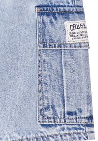 Tejido de mezclilla azul claro con un detalle de bolsillo, que presenta una etiqueta blanca cosida con el nombre de la marca y la información de fabricación.