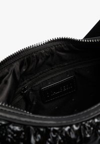 Interior de un bolso negro mostrando un bolsillo con cremallera y una etiqueta de marca de cuero cosida debajo de una cremallera parcialmente abierta.