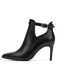 Botín de cuero negro hasta el tobillo con punta afilada, tacón alto de aguja y una correa ajustable con una hebilla dorada. Textura suave y diseño elegante.