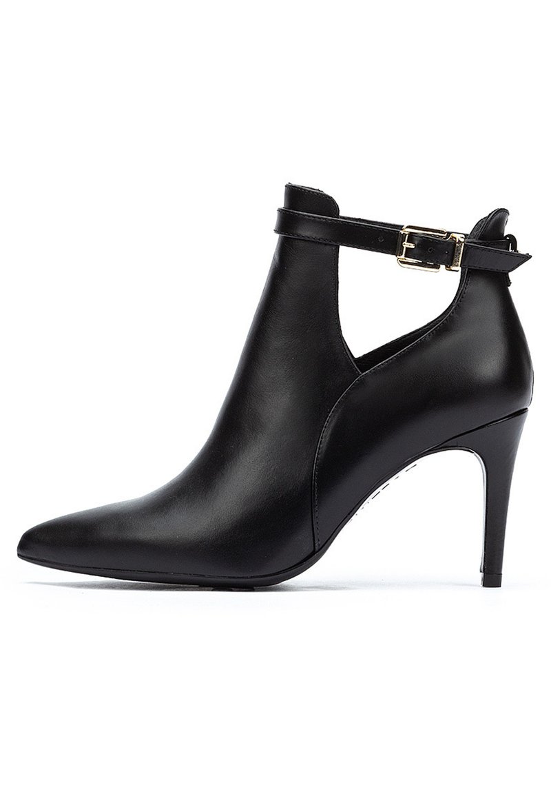 Botín de cuero negro hasta el tobillo con punta afilada, tacón alto de aguja y una correa ajustable con una hebilla dorada. Textura suave y diseño elegante.