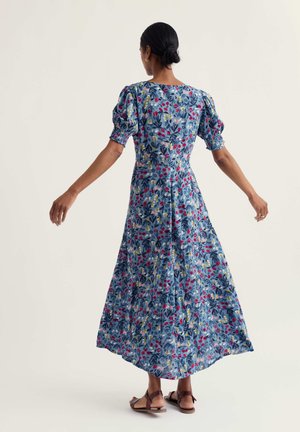 Kobieta o ciemnych włosach związanych w niski kok, ubrana w niebieską midi sukienkę w kwiaty z bufkami i brązowe sandały, stojąca z lekko wyciągniętymi ramionami.