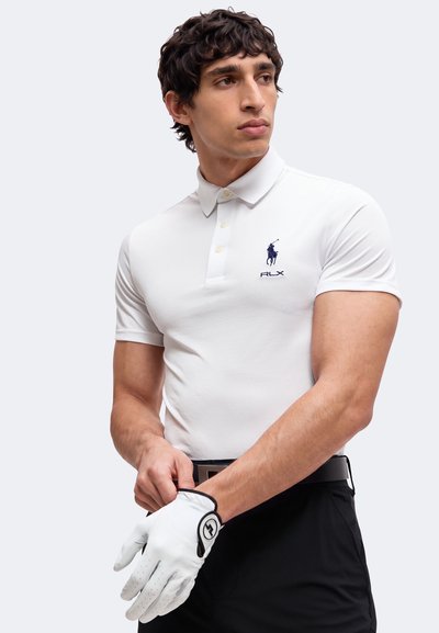 Homme ajustant un gant de golf blanc, portant un polo blanc avec un logo bleu et un pantalon noir sur un fond uni.