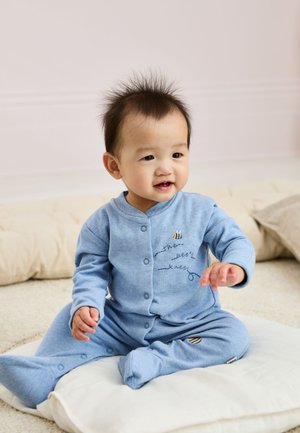 Bambino sorridente seduto su un cuscino bianco, indossa una tutina blu con ricamo a forma di ape, in un ambiente interno con luce soffusa.