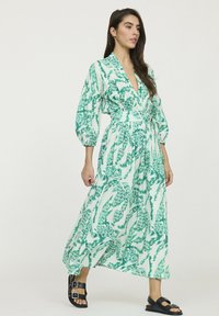 Robe maxi avec un décolleté en V, motif abstrait vert et blanc, manches amples, taille cintrée et jupe évasée, associée à des sandales noires.