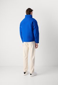 Giacca isolante blu con colletto alto, caratterizzata da una texture liscia. Indossata con pantaloni color crema e sneakers bianche con dettagli neri.