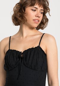 Vestido preto com um design texturizado, apresentando um decote em babados, alças ajustáveis e um pequeno detalhe de abertura frontal.