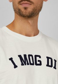 Weißes Baumwoll-T-Shirt mit Rundhalsausschnitt und dem Text "I MOG DI" in fetter, navyblauer Stickerei über der Brust. Glatte Textur, legerer Schnitt.