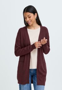 Cardigan bordeaux con apertura frontale, texture a maglia, maniche lunghe e tasche laterali, indossato su una maglietta beige chiaro a costine e jeans blu.