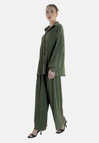 Ensemble deux pièces vert texturé comprenant une chemise à manches longues avec un col et un pantalon ample. Des sandales noires à lanières complètent le look.