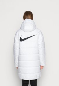 Personne portant une longue veste blanche rembourrée avec une capuche et un grand logo Nike noir dans le dos, se tenant devant un fond uni.