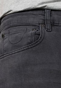 Jeans de mezclilla negra con un bolsillo visible, botón y detalles de costura. La textura del tejido es suave con un ligero brillo.