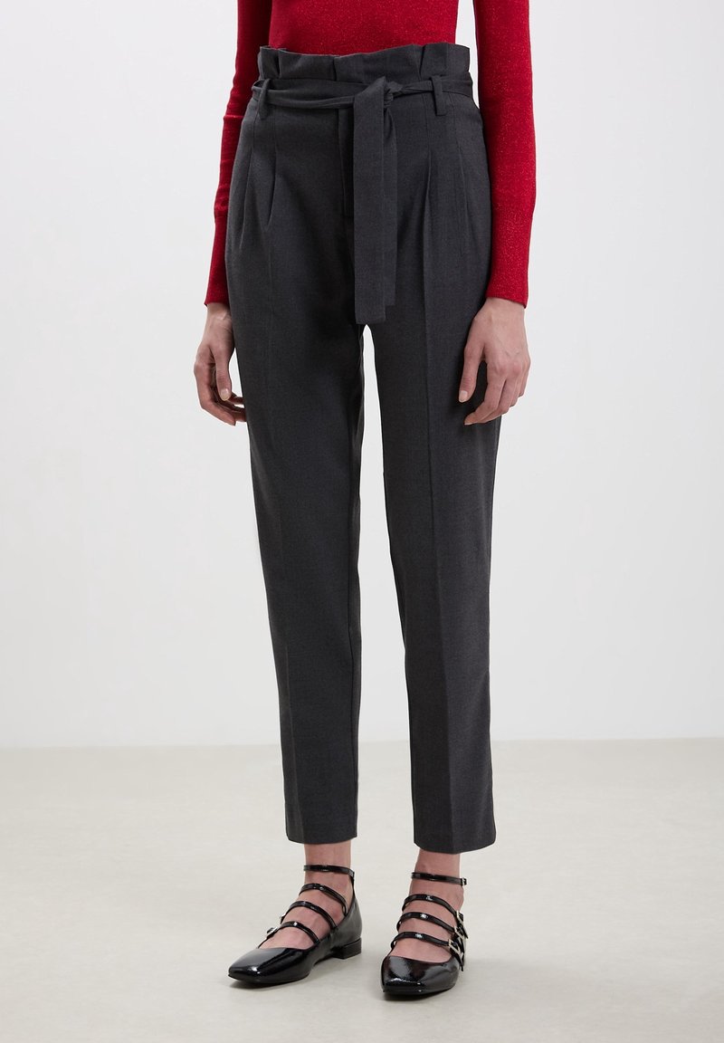 Femme portant un pantalon paperbag gris foncé taille haute avec ceinture nouée, un haut rouge à manches longues, et des chaussures plates noires à brides, debout sur un sol clair.
