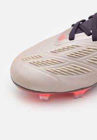 Close-up van een beige voetbalschoen met textuurpatronen, zwarte veters en rode noppen op de zool.