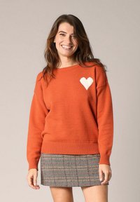 Pull en terre cuite avec un ourlet et des poignets côtelés, orné d'un motif de cœur blanc sur la poitrine. Porté avec une jupe à motif pied-de-poule.