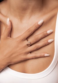 Goldener wellenförmiger Ring, der an einer Hand mit hellbeigen, polierten Nägeln getragen wird und auf einer nackten Schulter ruht. Die glatte Hautstruktur ist sichtbar.