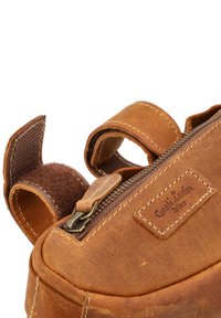 Brown leather bag with a textured surface, a zip closure, and two adjustable fabric straps. Includes a brand label stitched on the side.