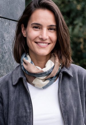 Une femme souriante aux cheveux bruns porte une écharpe à motifs et une veste grise en extérieur, près d’un mur en pierre.