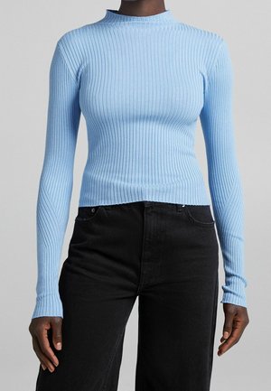 Femme portant un pull côtelé bleu clair ajusté à manches longues, associé à un jean noir taille haute sur un fond uni.