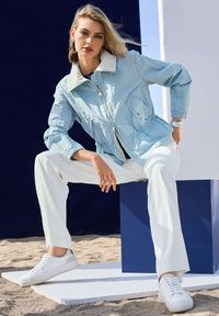 Lichtblauwe zip-up jack met een witte kraag, elastische manchetten en een trekkoord in de taille, gecombineerd met witte broeken en sneakers op zandgrond.